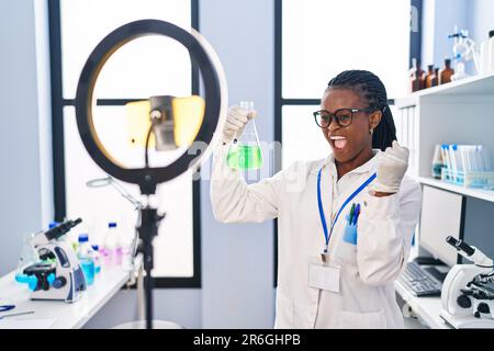 Donna africana con trecce che lavorano al laboratorio scienziato facendo tutorial con smartphone urlando orgoglioso, celebrando la vittoria e il successo molto eccitato Foto Stock