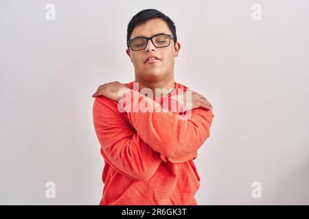 Giovane uomo ispanico con sindrome di Down in piedi su sfondo bianco abbracciarsi felice e positivo, sorridente fiducioso. amore di sé e cura di sé Foto Stock