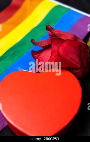 Un primo piano verticale di una scatola a forma di cuore e una rosa rossa vicino a una bandiera LGBT Foto Stock