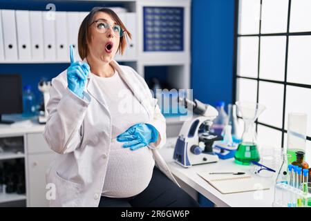 Donna incinta che lavora al laboratorio scienziato stupito e sorpreso guardando in su e indicando con le dita e le braccia sollevate. Foto Stock