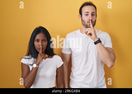 Coppia interrazziale in piedi su sfondo giallo chiedendo di essere tranquilla con il dito sulle labbra. silenzio e concetto segreto. Foto Stock