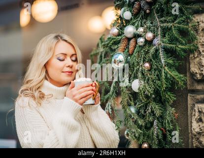 Ritratto all'aperto di felice bella donna rilassante su una terrazza di accogliente caffè o sala da tè, tenendo una tazza di cioccolata calda, indossando maglia bianca pullover Foto Stock