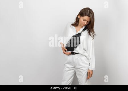 Donna bianca che indossa jeans bianchi, camicia e tracolla Borsa da cintura in pelle nera su sfondo grigio Foto Stock