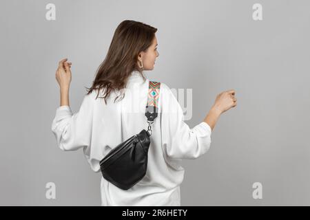 Donna bianca che indossa jeans bianchi, camicia e tracolla Borsa da cintura in pelle nera su sfondo grigio Foto Stock