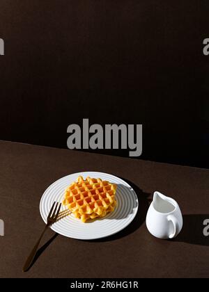 Un waffle appena fatto, servito su un piatto bianco con una forchetta d'argento e una tazza di caffè o tè, su uno sfondo bianco Foto Stock