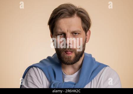 Giovane uomo caucasico che mostra la lingua rendendo i volti infantile alla macchina fotografica, imbrogliando intorno, scherzando, afilando con la faccia sciocca, brulicante, abuso di bullismo. Bel ragazzo isolato su studio sfondo beige interni Foto Stock