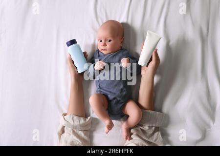 Madre che tiene crema corpo e polvere spolverante vicino al bambino piccolo cute sul letto, vista dall'alto Foto Stock