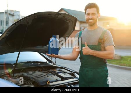 Un lavoratore sorridente che tiene un contenitore blu di olio motore e mostra i pollici in alto vicino all'auto all'aperto Foto Stock