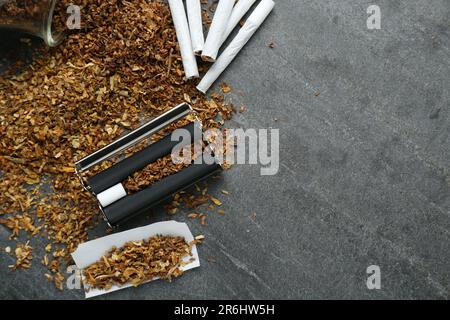 Composizione piatta con rullo e tabacco su tavolo grigio scuro, spazio per testo. Fare le sigarette arrotolate a mano Foto Stock