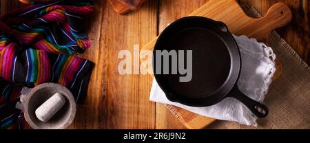 Padella vuota in ghisa sul tagliere. Sfondo cucina, colorato tradizionale tessuto messicano e piccola molcajete su rustico tavolo di legno. Panoram Foto Stock