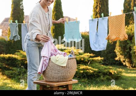 Donna sorridente appendere vestiti del bambino con i clothespins sulla linea di lavaggio per asciugare nel cortile posteriore, primo piano Foto Stock