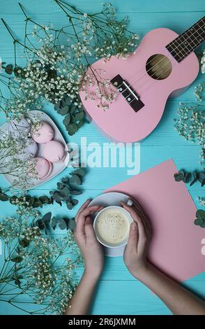 Ci sono ukulele rosa sul tavolo, la ragazza sta tenendo un bicchiere di caffè nelle sue mani. Ci sono fiori sul tavolo. C'è un piatto di dolci sul tavolo. Foto Stock