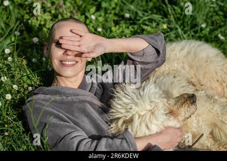 Gioiosa donna calva contenta con un cane è sdraiata sull'erba verde in una giornata di sole. Concetto di gioia di vita. Animale domestico. Foto Stock