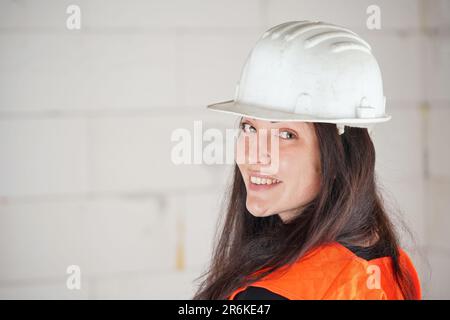 Giovane donna in elmetto bianco e giubbotto arancione ad alta visibilità, capelli lunghi e scuri, guardando sopra la spalla nella macchina fotografica, sorridendo. Costruzione sfocata si Foto Stock