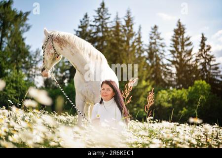 Donna seduta in una foresta primavera sole illuminato prato pieno di fiori margherite, bianco cavallo arabo accanto a lei Foto Stock