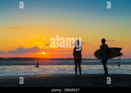 Surfista e donna fotografando il tramonto con smartphone sulla popolare spiaggia di Guiones, Playa Guiones, Nosara, Guanacaste, Costa Rica, America Centrale Foto Stock