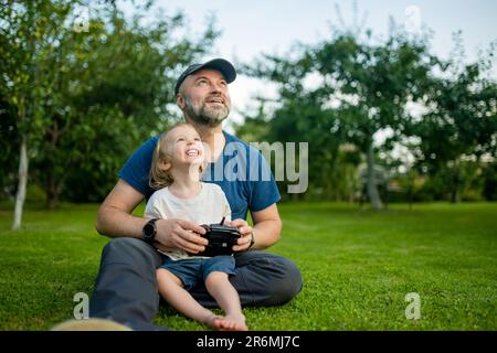 Padre e figlio guardano e navigano in un drone. Carino bambino ragazzo aiutando suo padre ad azionare un drone da telecomando. Tempo libero per tutta la famiglia. Foto Stock