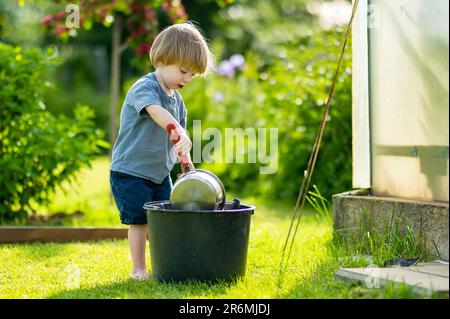 Carino biondo bambino piccolo giocare con una pentola di cottura all'aperto nel giardino. Bambino aiutando i genitori con il giardinaggio nel cortile in estate luminosa e soleggiata Foto Stock