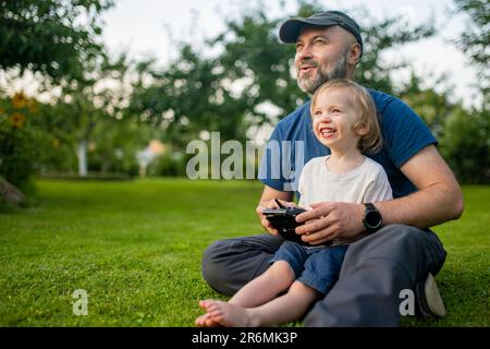 Padre e figlio guardano e navigano in un drone. Carino bambino ragazzo aiutando suo padre ad azionare un drone da telecomando. Tempo libero per tutta la famiglia. Foto Stock