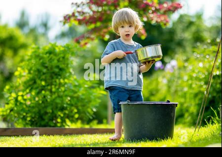 Carino biondo bambino piccolo giocare con una pentola di cottura all'aperto nel giardino. Bambino aiutando i genitori con il giardinaggio nel cortile in estate luminosa e soleggiata Foto Stock