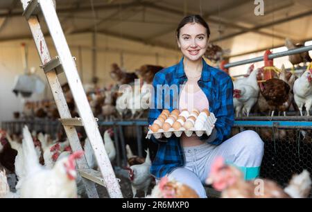 Giovane lavoratrice agricola europea che detiene una scatola di cartone piena di uova fresche di pollo nell'azienda avicola Foto Stock