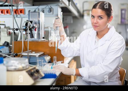 Biologo giovane intelligente in camice bianco con pipetta meccanica da laboratorio per la miscelazione di prodotti chimici in laboratori moderni Foto Stock