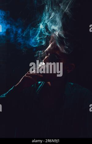Nero giovane uomo con occhiali Fumante Cigar con nebbia su sfondo nero, luce chiave bassa. Foto Stock