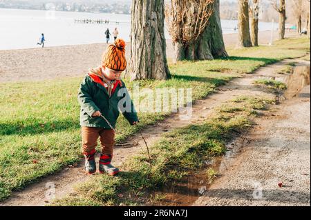 Carino bambino ragazzo giocare con pudle di fango all'esterno, indossare giacca calda e stivali invernali, felice infanzia Foto Stock