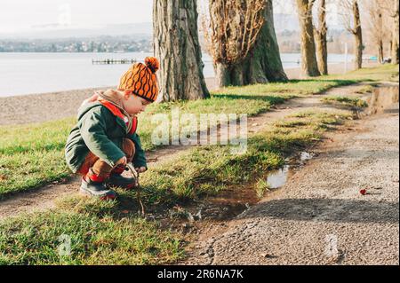 Carino bambino ragazzo giocare con pudle di fango all'esterno, indossare giacca calda e stivali invernali, felice infanzia Foto Stock