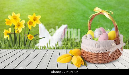 Coniglietto che si nasconde in erba verde vicino al ponte di legno con cestino di vimini pieno di uova di Pasqua Foto Stock