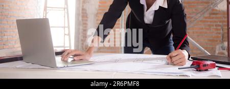 Giovane architetto caucasico donna alla ricerca di un computer portatile e di un piano di disegno di lavoro per la pianificazione di una casa di costruzione sulla scrivania, ingegnere pensare e disegnare blu Foto Stock