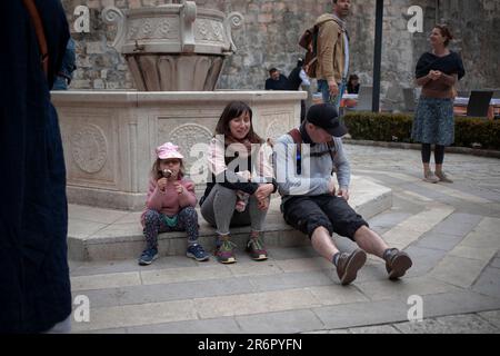 Cattaro, Montenegro, 13 aprile 2023: Una coppia turistica con una figlia che prende una pausa seduto sulla piazza accanto a una fontana di acqua potabile Foto Stock