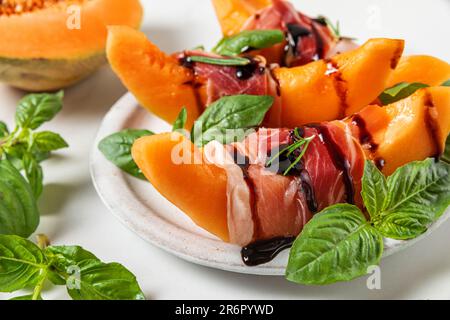Prosciutto con fettine di melone cantaloupe, salsa all'aceto balsamico e basilico in un piatto su fondo bianco Foto Stock