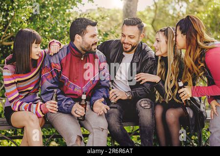 Un gruppo di amici, due uomini e tre donne, seduti insieme su una panchina del parco, per una conversazione informale e per tenere bevande in bottiglia. Lo sono Foto Stock