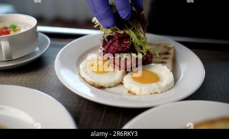 Rukua cosparge la lattuga sulle uova strapazzate su un piatto. Uova fritte con salsicce e insalata al mattino per colazione. Mani maschili con forchetta e. Foto Stock