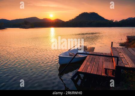 Splendida vista del tramonto che splende sulla montagna e sul lago con barca al molo di legno nella scena rurale Foto Stock