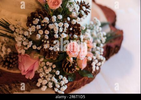 Una torta nuziale bianca a tre livelli adornata con rose rosa, coni di pino e piccoli fiori bianchi e rosa Foto Stock