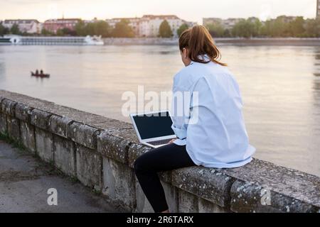 Donna che lavora in remoto sul suo computer portatile fuori dall'ufficio seduto sul lungomare di fronte al fiume città europea. Foto Stock