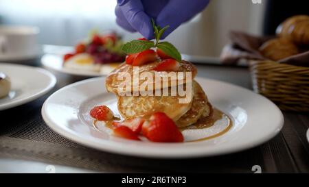 Primo piano di una mano che mette una foglia di menta sui pancake. Lo chef prepara la colazione in camera. Le mani del cuoco mettono le foglie di menta sui pancake. Foto Stock