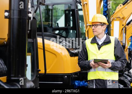 Ingegnere in un casco con un tablet digitale stand accanto agli escavatori da cantiere Foto Stock