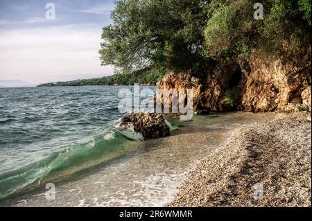 Sulla costa del Peloponneso, piccole onde di acqua cristallina si rompono su una romantica spiaggia con rocce e boschi sullo sfondo Foto Stock