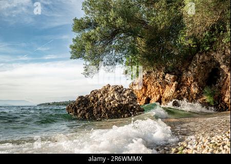 Sulla costa del Peloponneso, piccole onde di acqua cristallina si rompono su una romantica spiaggia con rocce e boschi sullo sfondo Foto Stock