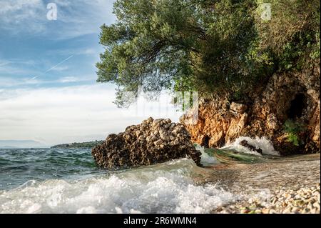 Sulla costa del Peloponneso, piccole onde di acqua cristallina si rompono su una romantica spiaggia con rocce e boschi sullo sfondo Foto Stock