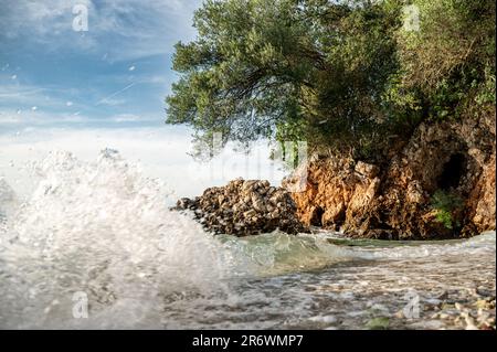 Sulla costa del Peloponneso, piccole onde di acqua cristallina si rompono su una romantica spiaggia con rocce e boschi sullo sfondo Foto Stock