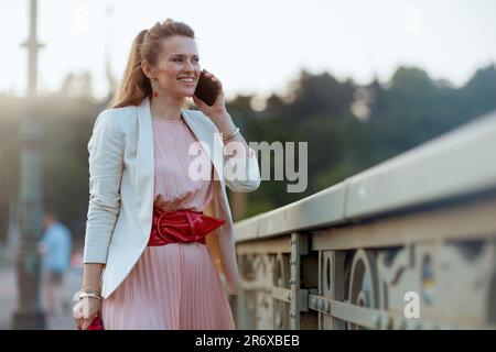 donna sorridente elegante di 40 anni in abito rosa e giacca bianca in città che parla su uno smartphone sul ponte. Foto Stock