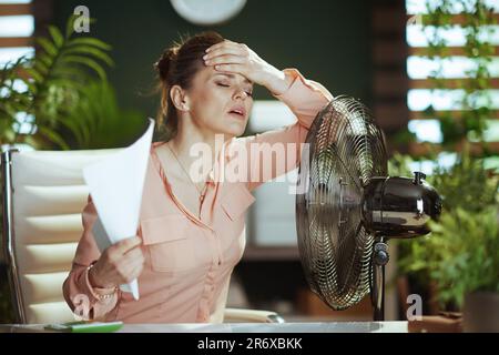 Ambiente di lavoro sostenibile. moderna dipendente di 40 anni al lavoro con documenti e ventilatore elettrico che soffre di calore estivo. Foto Stock