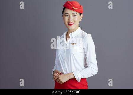 elegante donna asiatica sorridente in gonna rossa e cappello uniforme isolato su sfondo grigio. Foto Stock