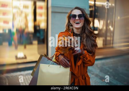 Ciao settembre. donna sorridente di mezza età in trench marrone con borse per la spesa e foglie gialle autunnali vicino al negozio in città.