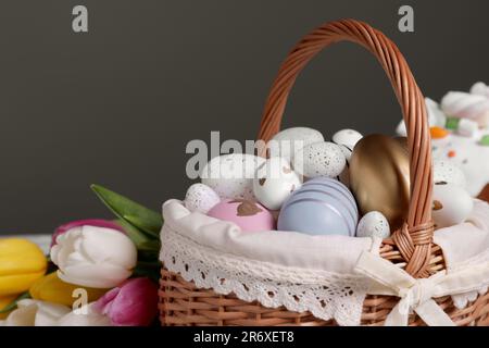 Cestino di vimini con uova di Pasqua decorate in modo festivo e bellissimi tulipani su sfondo grigio scuro, primo piano Foto Stock