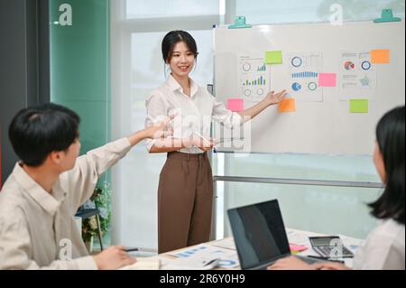 Una giovane donna d'affari asiatica bella e fiduciosa o una assistente finanziario femminile sta spiegando e presentando il suo rapporto grafico finanziario al suo team Foto Stock